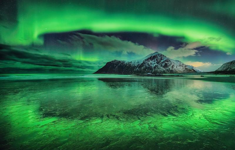 Image of a snow-capped mountain next to a wide expanse of frozen water, with a vivid aurora making almost an arc in the sky from left to right, which is reflected in the ice below