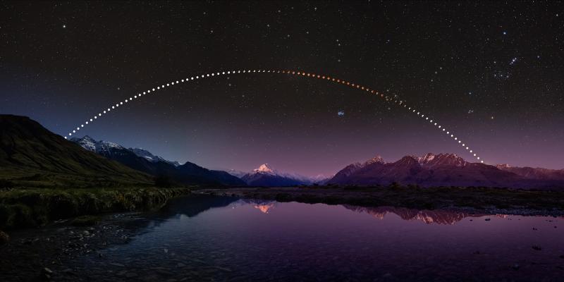 Image of a lake at twilight with snow-capped mountains behind it, while in the sky multiple images of a lunar eclipse have been compiled to make an arc showing its movement through the sky and its colour change from white to red, back to white