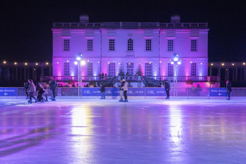 Image of the Queen's House Ice Rink with the classical building of the Queen's House in the background, lit up purple and blue, with a quite empty rink in front of it, only around 8 people on it
