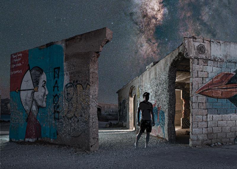 A night sky photograph of the Milky Way taken amongst ruined buildings. The eerie scene features graffiti on crumbling walls, with the figure of a man in shadow standing by a painting of a bird. A stream of stars bisects the sky above them