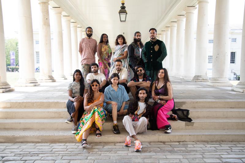 A photo of 13 South Asian creatives some of whom are sitting and some are standing in the colonnades next to the Queen's House. A central lamp hangs above the group and they are framed by the white stone columns.  