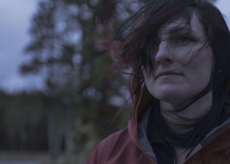 A young woman photographed in a forest at dusk. She is looking pensively beyond the camera, with her hair blowing across her face in the wind 