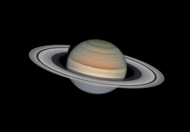 An image of Saturn taken using a telescope. The rings of the planet are clearly visible round a dusky yellow and orange globe. The rest of the frame is pitch black