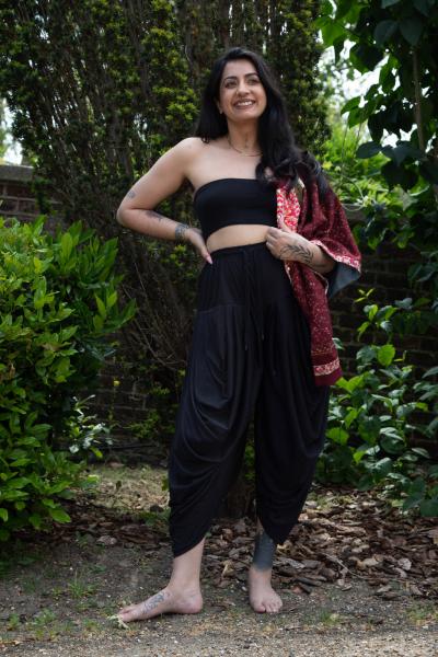Portrait photograph of a woman standing bare-footed in a wood. She wears loose black trousers and a black crop top with a patterned silk jacket slung over her left shoulder