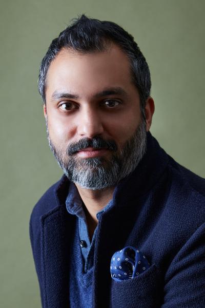 A headshot photo of Ajesh Patalay, who looks directly at the camera. He wears a blue shirt and navy jacket and has short black hair with a full moustache and beard with grey speckles.