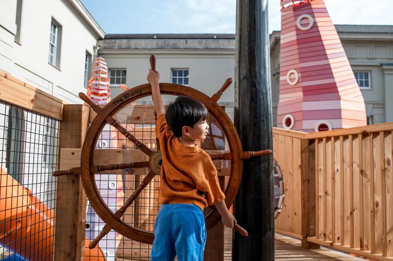 A young boy takes the helm of a large ship's wheel, part of an outdoor playground apparatus at the National Maritime Museum
