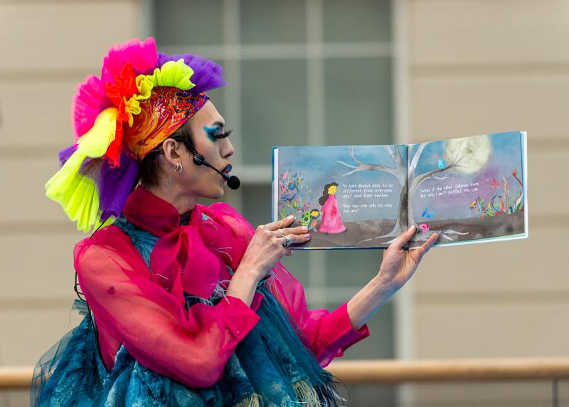 A drag performer with brightly coloured wig and nautical inspired dress reads from a picture book
