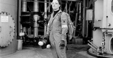 A black and white photo of an oil worker. She wears glasses and overalls, and stands looking at the camera in front of rig pipes