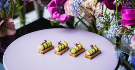 Moving venue catering's Courgette cannelloni, cashew curd with multigrain croute on a white plate next to pink and purple table flower centerpiece