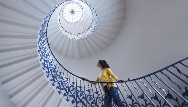 A woman stands looking up at the spiral staircase in the Queen's House known as the Tulip Stairs