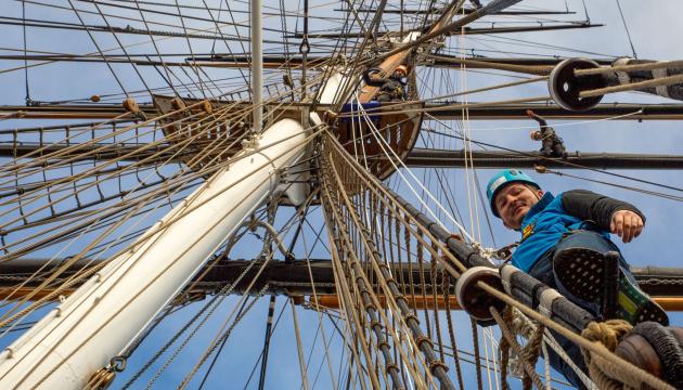 A photography looking up at the rigging of sailing ship Cutty Sark. A man wearing a helmet and harness is standing in the ropes looking down at the photographer