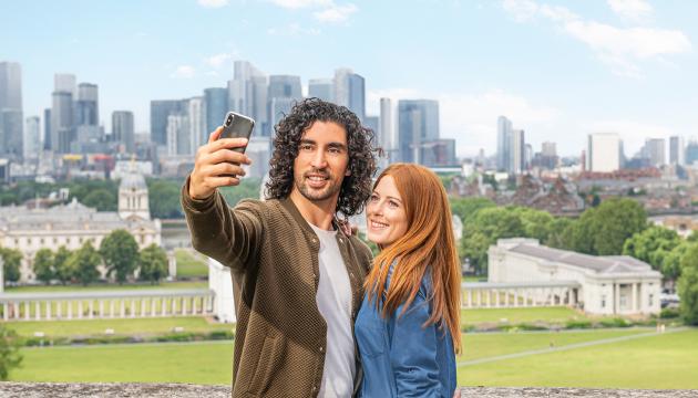 A couple takes a selfie at the top of Greenwich Park, with the River Thames and Canary Wharf in the background