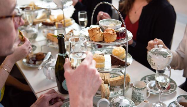A table laid out for afternoon tea, with people gathered round a platter of sandwiches, scones and cakes, plus bottles of prosecco and pots of tea